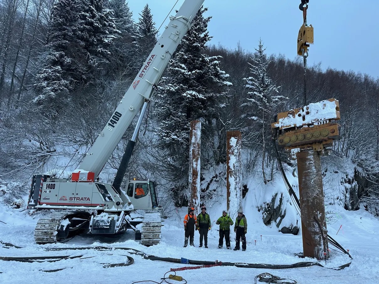 Crane and No Shock connected to a vibratory hammer beside steel piles on a snow-covered jobsite